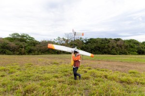 A man in an orange jacket, black pants, and white bucket hat faces the left side of the frame, carrying a fixed-wing uncrewed aircraft over his shoulder across a field of green grass. On one wing is the words Black Swift Technologies in a serif font with a silhouette of a black bird. On the other wing is the NASA logo: a blue circle with a red vector and the word NASA in white text.