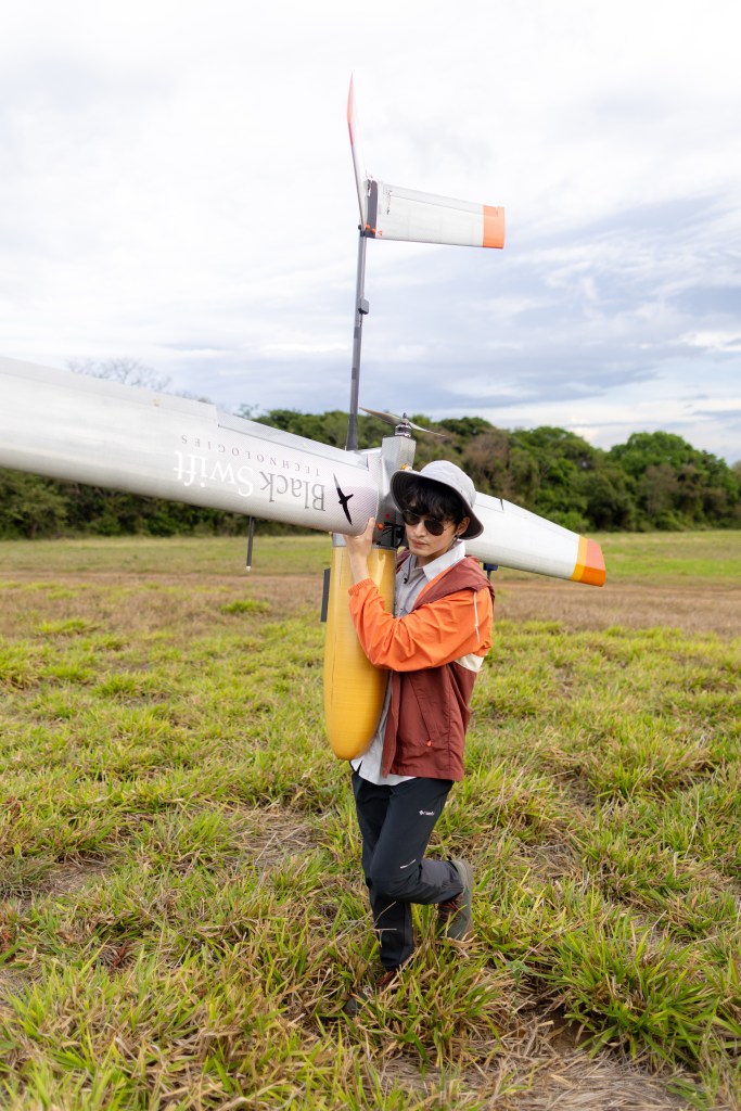 A man in an orange jacket, black pants, and white bucket hat faces the left side of the frame, carrying a fixed-wing uncrewed aircraft over his shoulder across a field of green grass. On the wing reaching towards the camera is the words Black Swift Technologies in a serif font with a silhouette of a black bird.