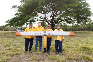 Four men in yellow long sleeve shirts stand in a grassy field, holding a silver fixed-wing uncrewed aircraft with a yellow nose cone pointed towards the ground. In the background is a large tree.