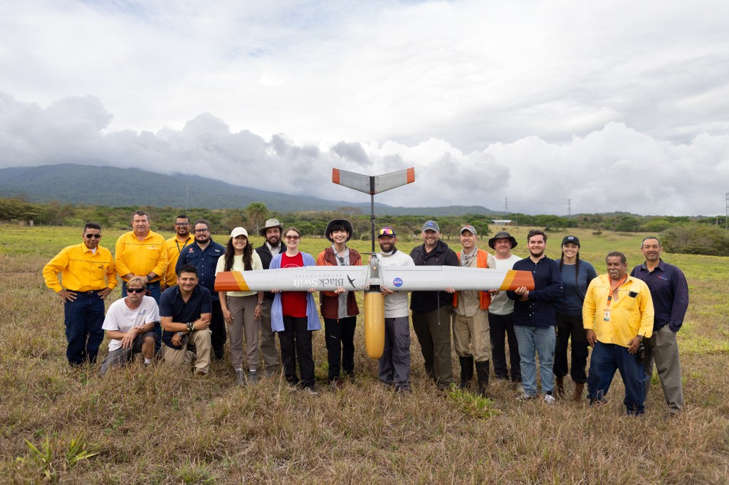 A large group of people pose in a grassy field, holding a silver fixed-wing uncrewed aircraft with a yellow nose cone pointed towards the ground. In the background is a blueish mountain and cloudy skies.