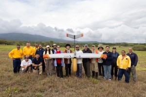 A large group of people pose in a grassy field, holding a silver fixed-wing uncrewed aircraft with a yellow nose cone pointed towards the ground. In the background is a blueish mountain and cloudy skies.
