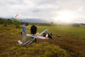 Two men work on a small gray fixed-wing aircraft mounted on a sliver launcher shaped like a tripod, pointing to the right of the frame. They stand in a red and green grassy field, with a blueish mountain and cloudy skies with the sun breaking through just above the horizon in the distance.
