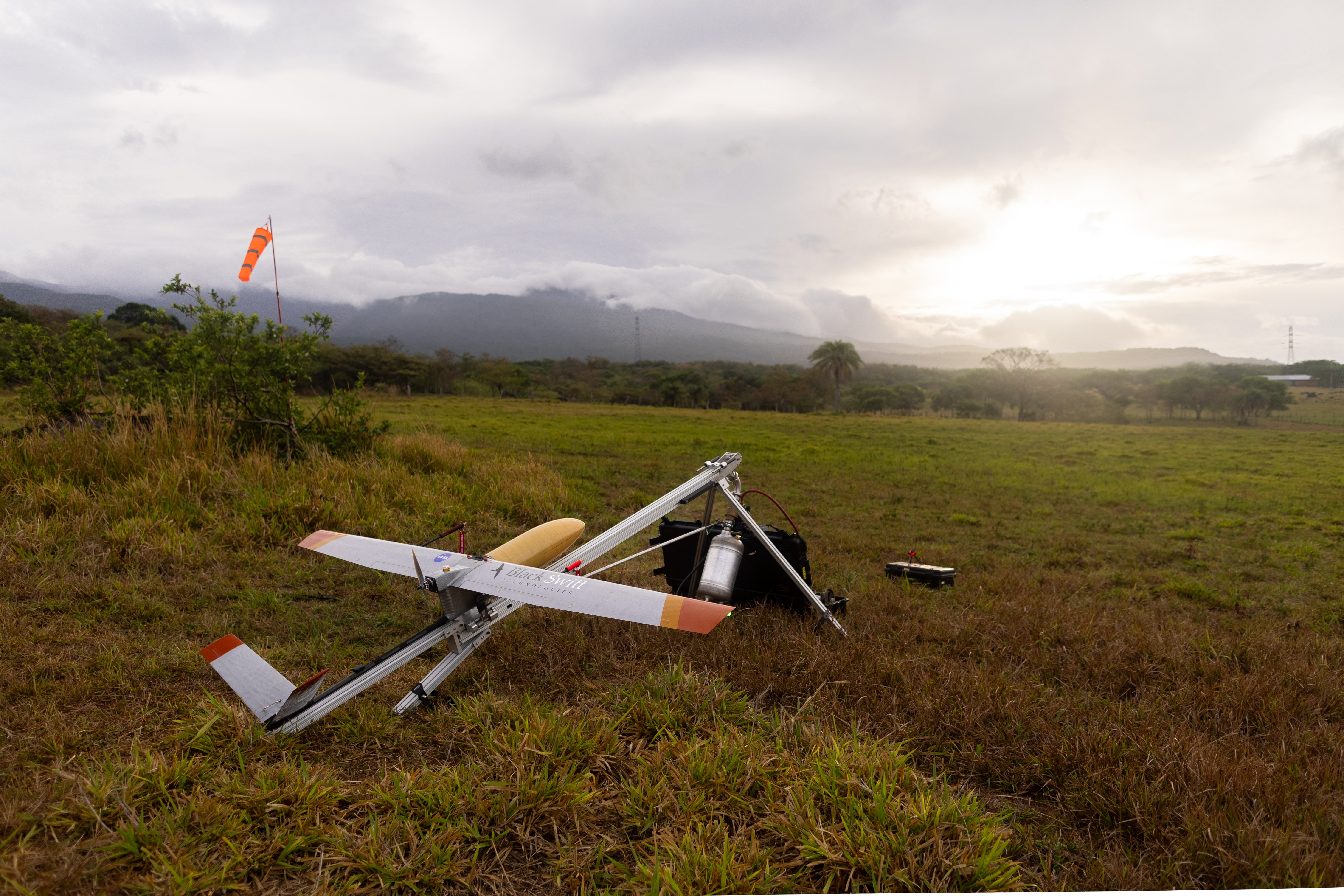 A small gray fixed-wing aircraft with a yellow nose cone sits mounted on a sliver launcher shaped like a tripod, pointing to the right of the frame. They stand in a red and green grassy field, with a blueish mountain and cloudy skies with the sun breaking through just above the horizon in the distance.