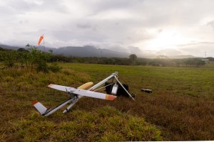 A small gray fixed-wing aircraft with a yellow nose cone sits mounted on a sliver launcher shaped like a tripod, pointing to the right of the frame. They stand in a red and green grassy field, with a blueish mountain and cloudy skies with the sun breaking through just above the horizon in the distance.