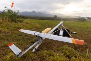 A small gray fixed-wing aircraft with a yellow nose cone sits mounted on a sliver launcher shaped like a tripod, pointing to the right of the frame. They stand in a red and green grassy field, with a blueish mountain and cloudy skies with the sun breaking through just above the horizon in the distance.