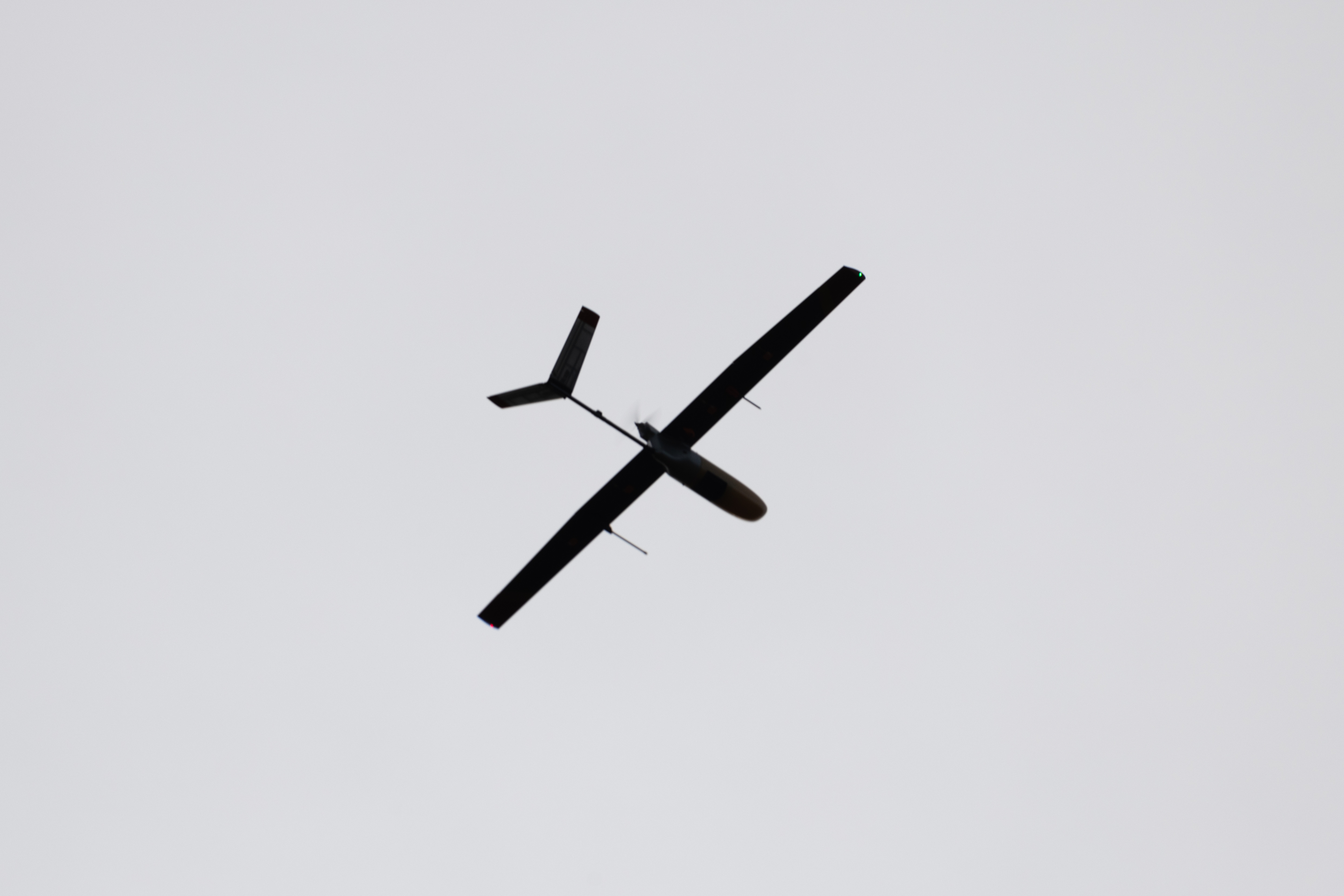 The small black shape of an uncrewed fixed-wing aircraft is silhouetted against gray clouds with the sun breaking through, flying from right to left.