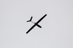 The small black shape of an uncrewed fixed-wing aircraft is silhouetted against gray clouds with the sun breaking through, flying from right to left.