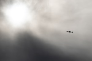 The small black shape of an uncrewed fixed-wing aircraft is silhouetted against gray clouds with the sun breaking through, flying from right to left.