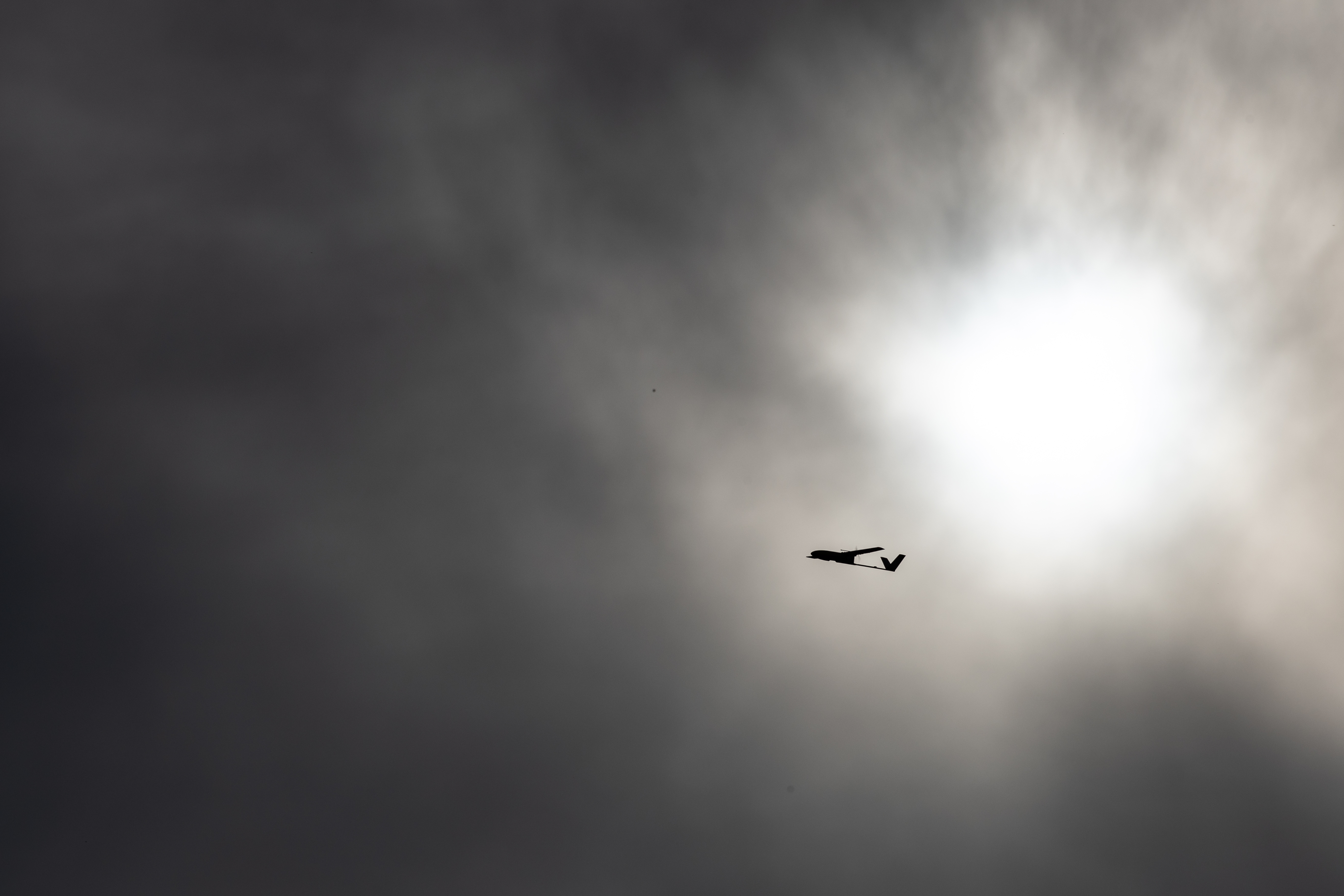 The small black shape of an uncrewed fixed-wing aircraft is silhouetted against gray clouds with the sun breaking through, flying from right to left.