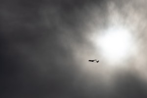 The small black shape of an uncrewed fixed-wing aircraft is silhouetted against gray clouds with the sun breaking through, flying from right to left.