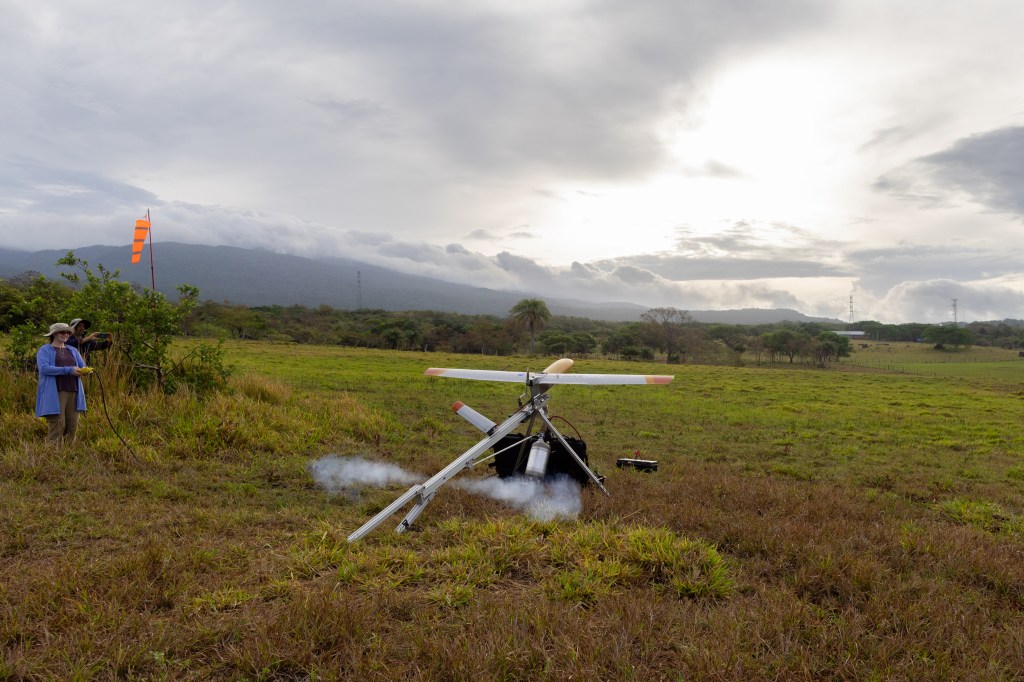 A woman in a periwinkle jacket and black pants stands in a field of tan and green grass, watching a small gray fixed-wing aircraft with a yellow nose cone as it flies off of a silver launcher. Two puffs of white vapor vent out of the back of the launcher. In the background is a blueish mountain and cloudy skies.