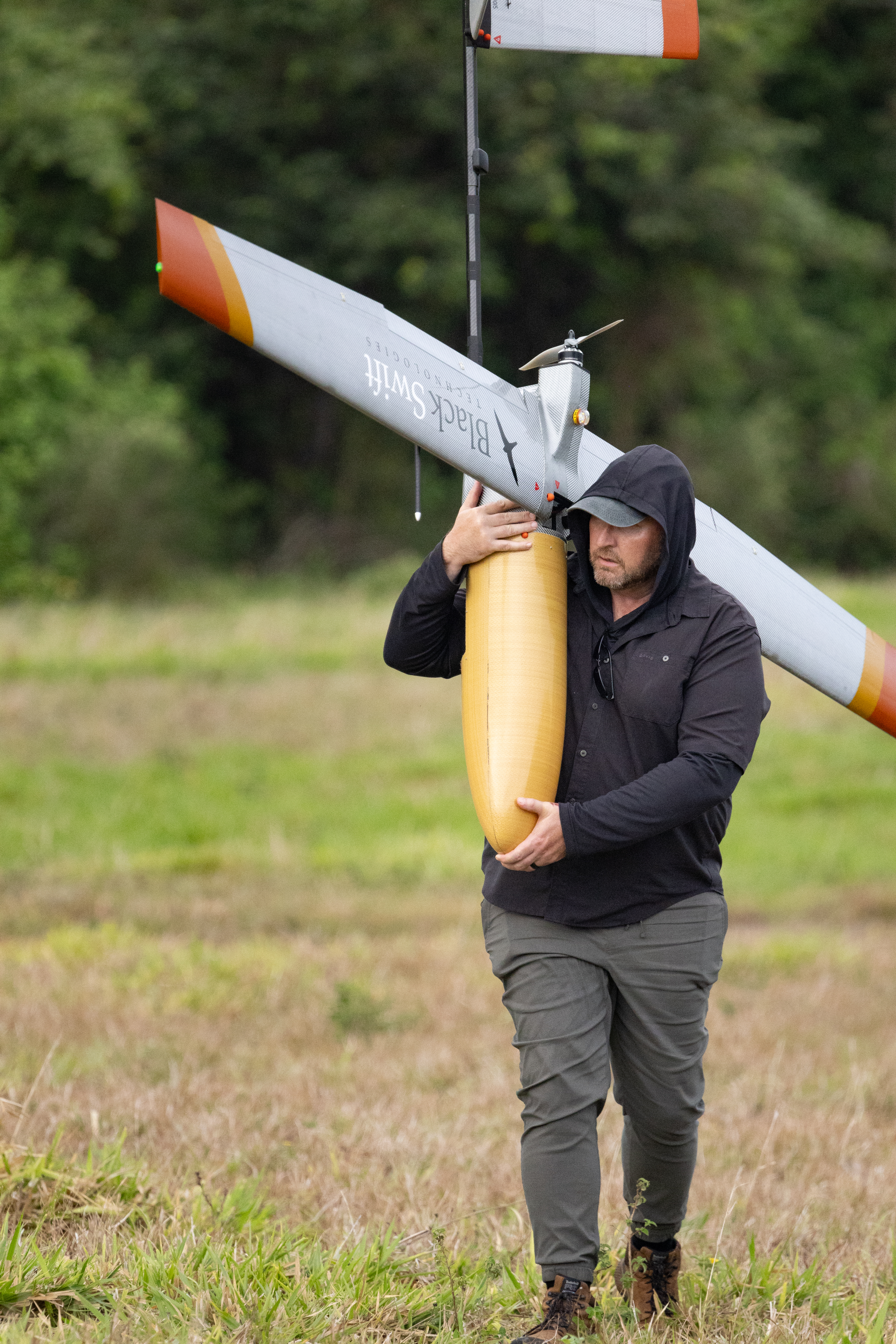 A man in a black hooded jacket faces the left side of the frame, carrying a fixed-wing uncrewed aircraft over his shoulder across a field of green and tan grass. On the wing reaching towards the camera is the words Black Swift Technologies in a serif font with a silhouette of a black bird.