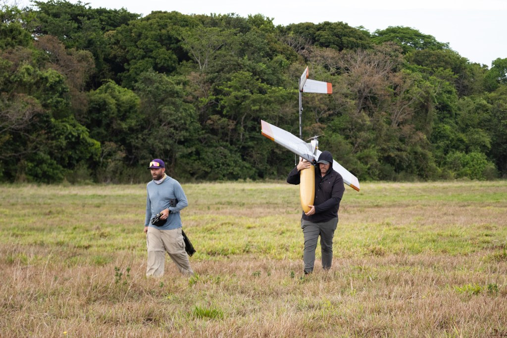 A man in a black hooded jacket carries a fixed-wing uncrewed aircraft over his shoulder across a field of green and tan grass, while a man in a blue long sleeve walks in front of him. In the background is a stretch of bright green trees.