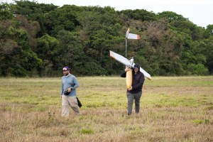 A man in a black hooded jacket carries a fixed-wing uncrewed aircraft over his shoulder across a field of green and tan grass, while a man in a blue long sleeve walks in front of him. In the background is a stretch of bright green trees.