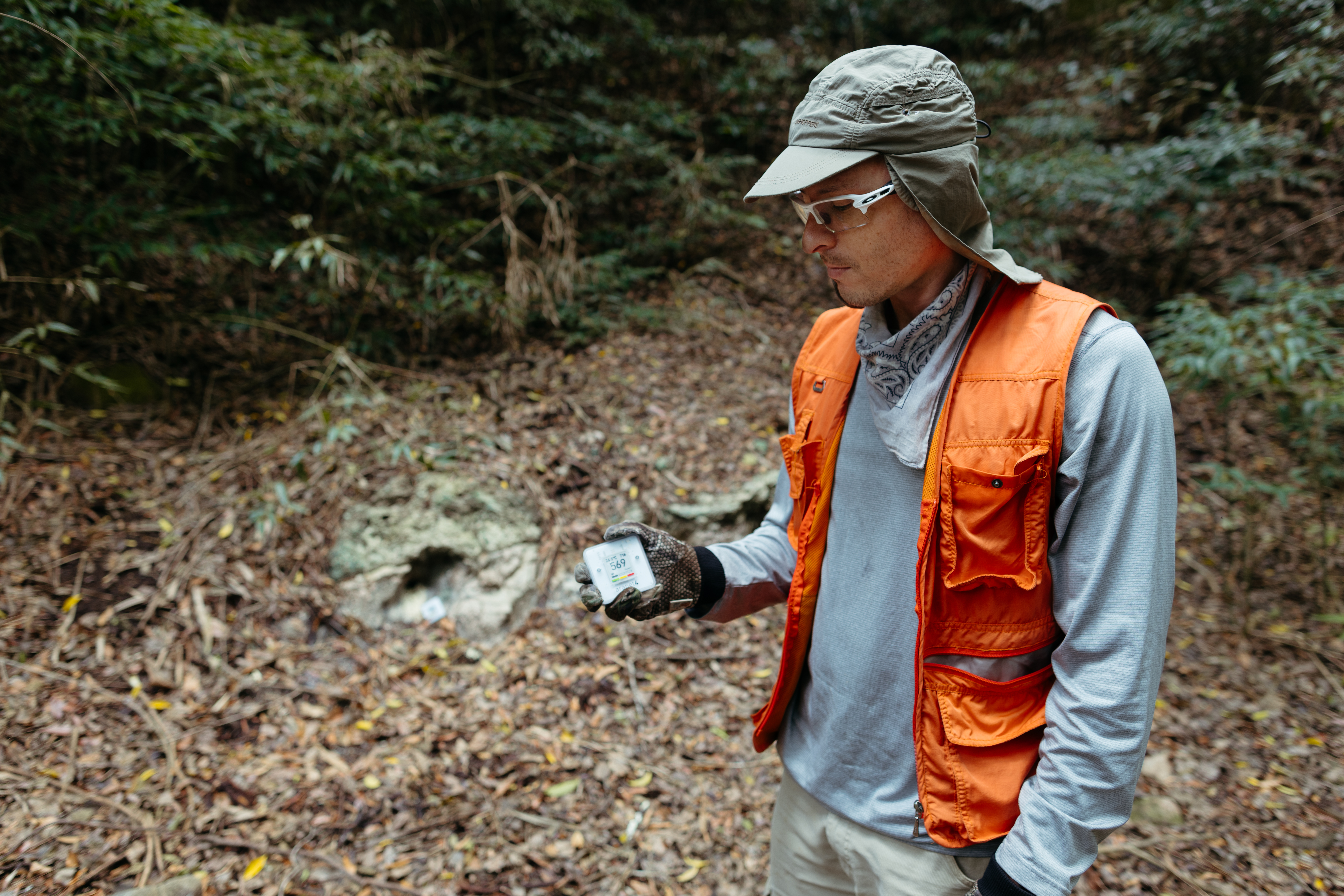 A man in a blue long sleeve, orange vest, and khaki green hat looks down at a small white box in his right hand. In the background is a hillside covered in leaves, with a small hole.