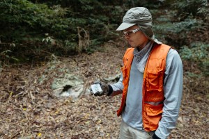 A man in a blue long sleeve, orange vest, and khaki green hat looks down at a small white box in his right hand. In the background is a hillside covered in leaves, with a small hole.