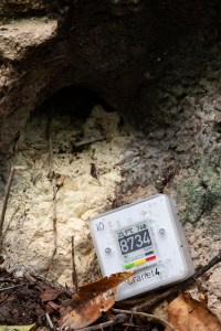 A small white box with a digital screen showing the number 8734 leans against a tan rock, with a dark hole in the top of the frame.