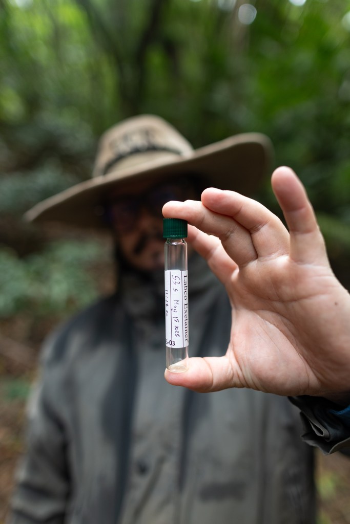 A man in a khaki green jacket and large hat holds a small vial towards the camera, with a handwritten label.