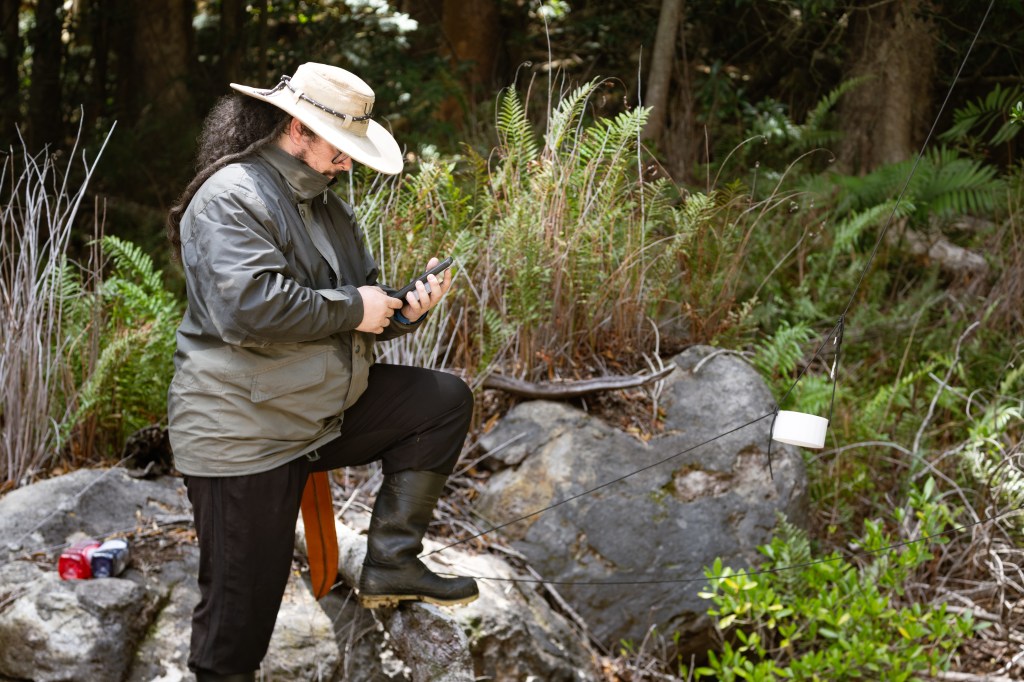 A man in a khaki green jacket and large hat stands with his foot up on a rock, checking readouts on his phone. In front of him, a round white box hangs on a black string.