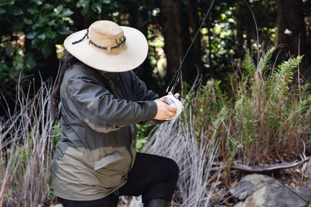 A man in a khaki green jacket and large hat stands with his foot up on a rock, working on a round white box hangs on a black string.