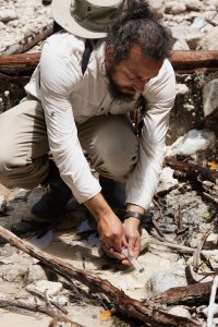 A man with long black hair and a black beard, wearing a white long sleeve, crouches over a shallow stream and takes a sample with a plastic syringe.