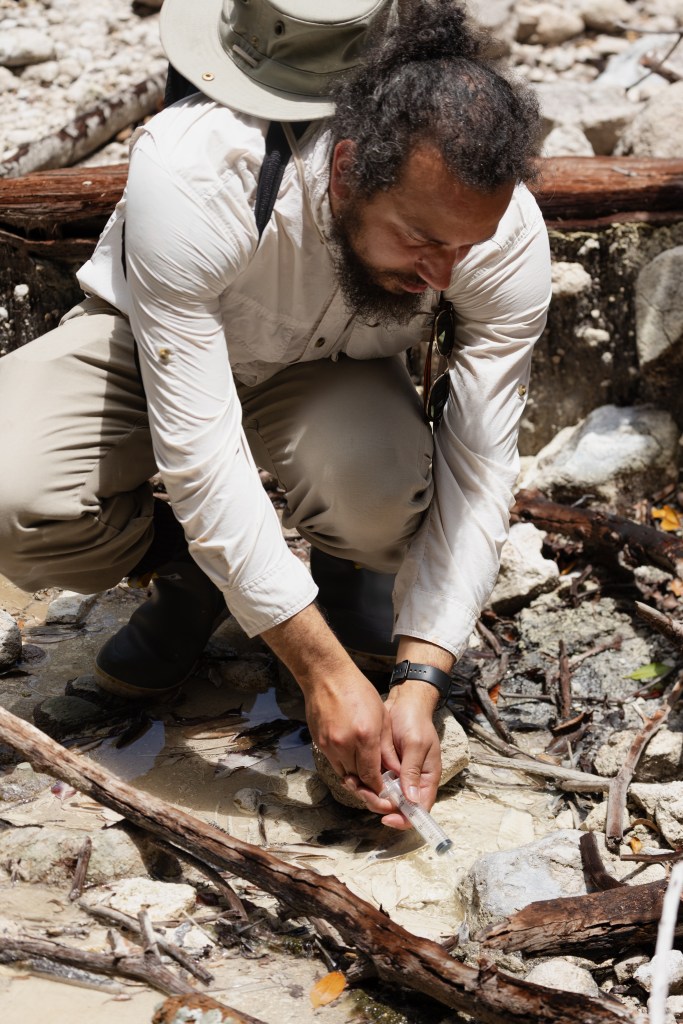 A man with long black hair and a black beard, wearing a white long sleeve, crouches over a shallow stream and takes a sample with a plastic syringe.