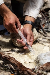 A close-up shot of a man's hands, wearing a black watch, using a plastic syringe over a very shallow stream.