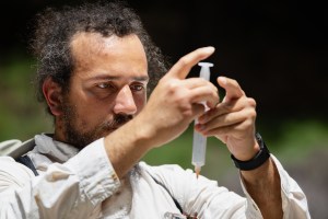 A man with long black hair and a black beard, wearing a white long sleeve, holds up a plastic syringe.