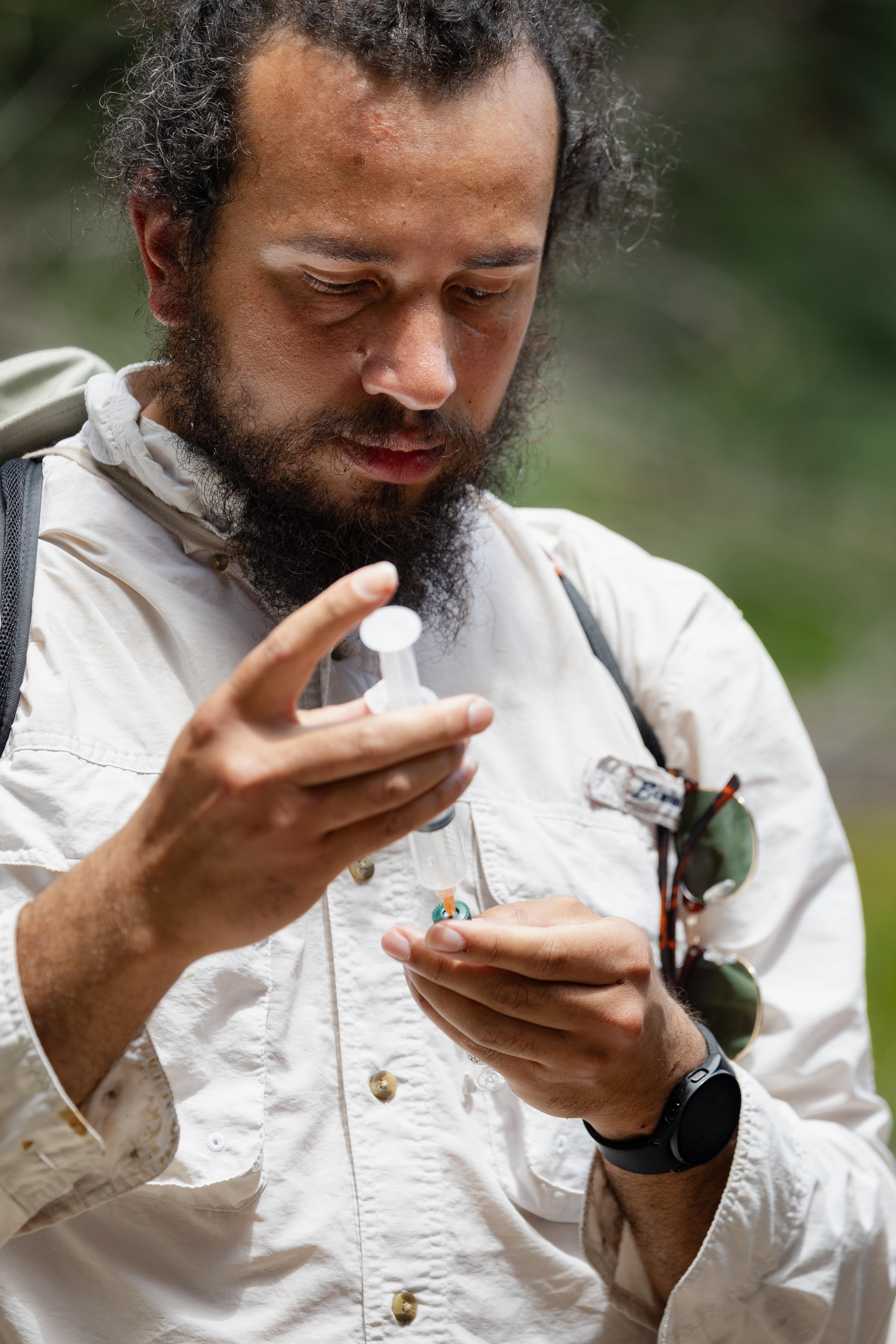 A man with long black hair and a black beard, wearing a white long sleeve, looks down at a plastic syringe in his hands.