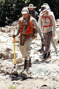 Four men in hats, boots, and hiking clothes walk along white rocks towards the camera.