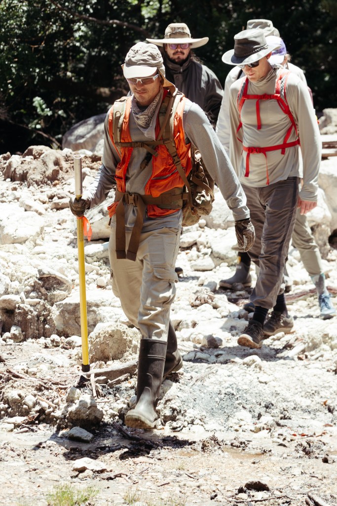 Four men in hats, boots, and hiking clothes walk along white rocks towards the camera.