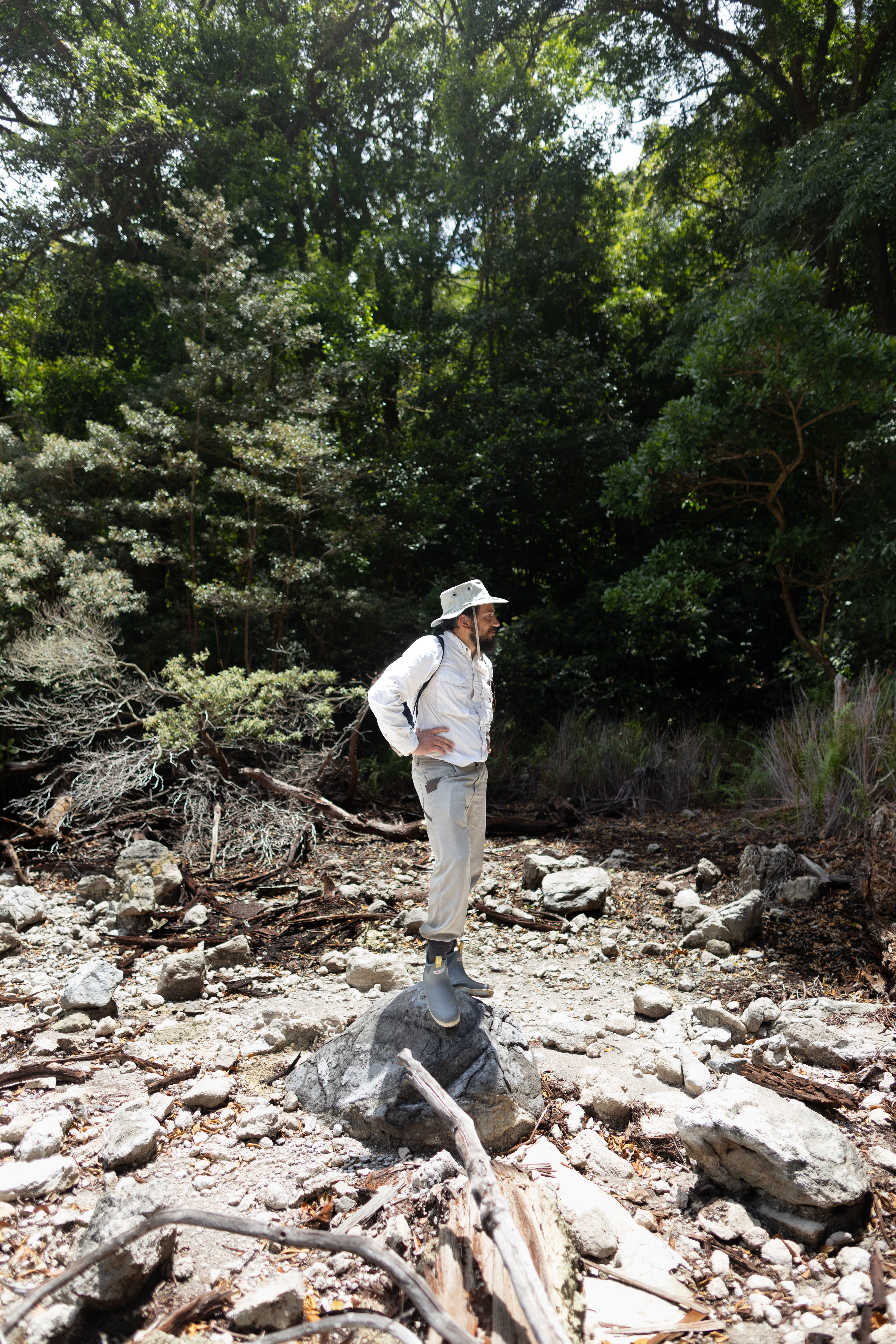 A man in a white shirt with tan pants and a tan hat stands on a rock, looking to the right of the frame. In the background is a green forest.