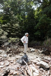 A man in a white shirt with tan pants and a tan hat stands on a rock, looking to the right of the frame. In the background is a green forest.