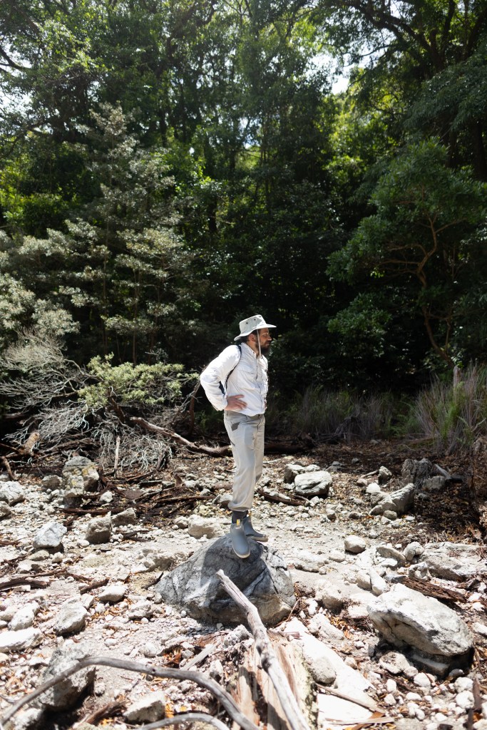 A man in a white shirt with tan pants and a tan hat stands on a rock, looking to the right of the frame. In the background is a green forest.