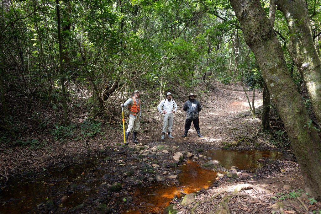 Three men in hiking attire pose in a forest next to a reddish-orange stream.