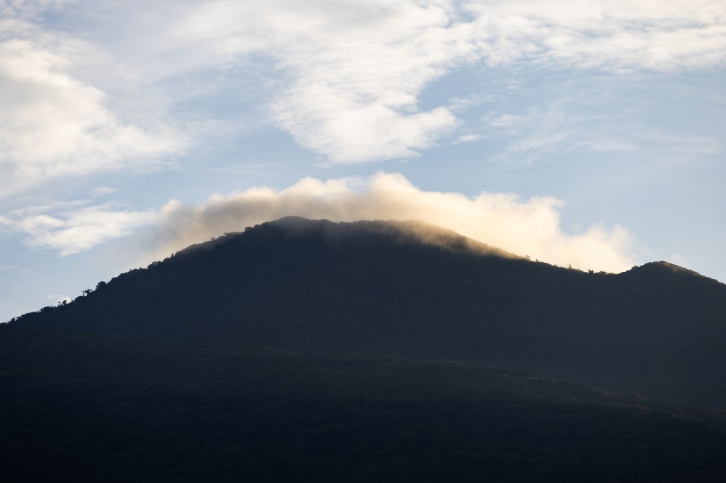 A dark blue mountain is partially silhouetted against a blue sky dotted in clouds. A few clouds sit right around the summit.