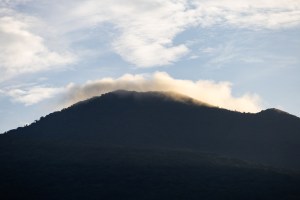 A dark blue mountain is partially silhouetted against a blue sky dotted in clouds. A few clouds sit right around the summit.