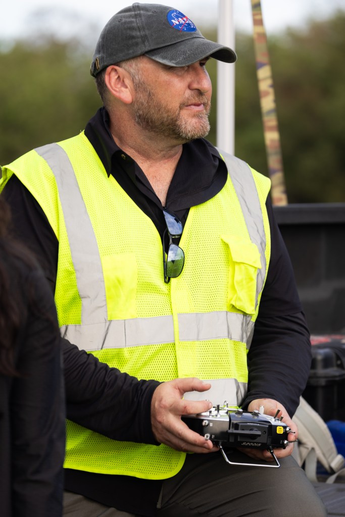 A man with gray and brown scruff, wearing a black long sleeve, a neon yellow high-vis vest, and a dark gray baseball hat with a NASA logo, holds a drone controller and looks off to the right of the frame.