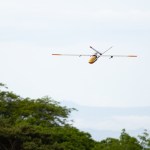 A gray uncrewed fixed-wing aircraft with a yellow nose cone flies towards the left of the frame against a cloudy sky, with the tops of trees in the lower third of the image.