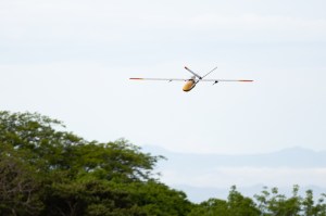 A gray uncrewed fixed-wing aircraft with a yellow nose cone flies towards the left of the frame against a cloudy sky, with the tops of trees in the lower third of the image.