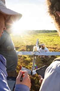 The camera shoots through two people looking at each other and just visible on the edges of the frame. In the center, a man crouches next to a gray uncrewed fixed-wing aircraft with a yellow nose cone.