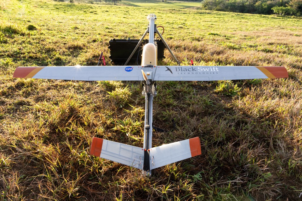 A gray uncrewed fixed-wing aircraft with a yellow nose cone at orange-tipped wings sits on a tripod launcher in a field of green and tan grass.