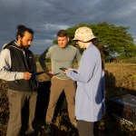 Three people in hiking clothes stand together in a field of tan grass, looking down at tablets. Behind them is a dark cloudy sky.