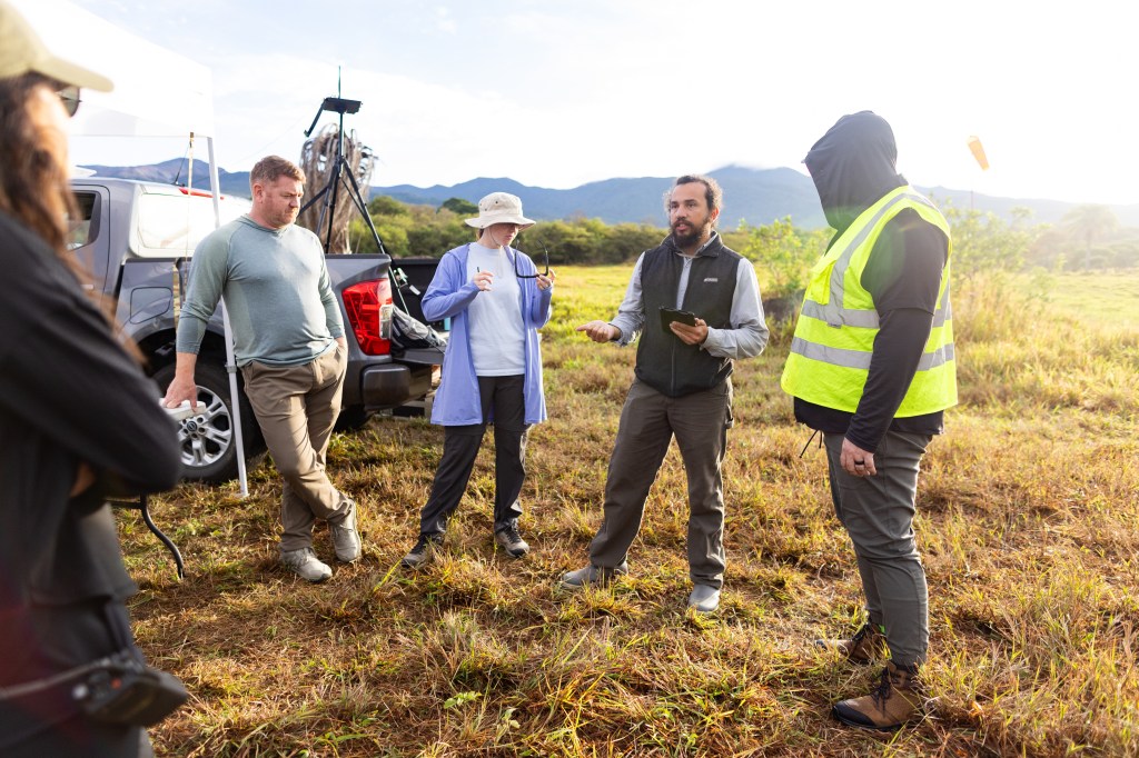 A man with long dark hair speaks to a group of people gathered in a gold field of grass.