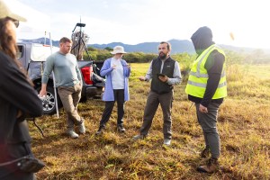 A man with long dark hair speaks to a group of people gathered in a gold field of grass.