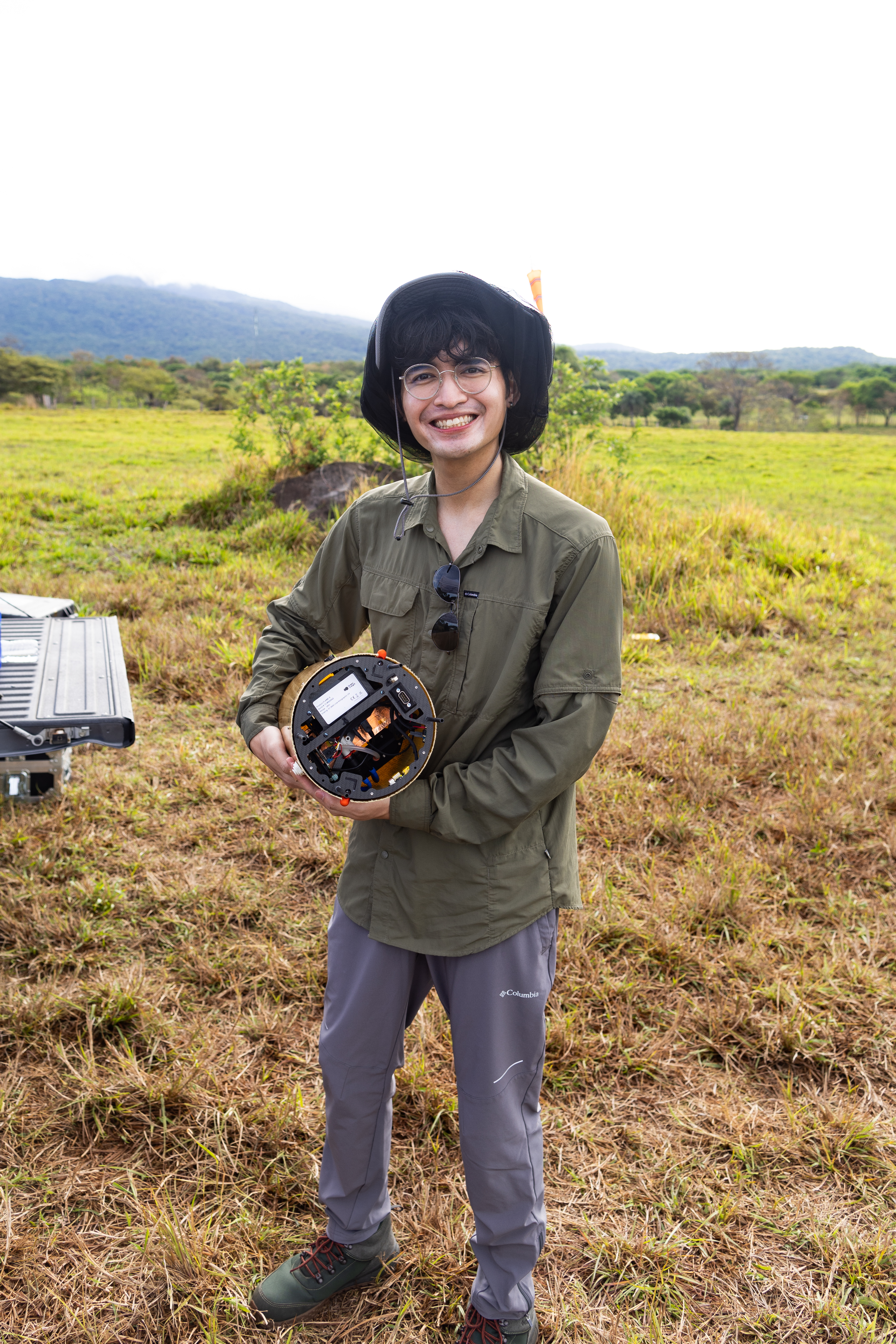 A man in an olive green longsleeve and black bucket hat smiles at the camera while holding a cylindrical device. He stands in a field of grass with a blue mountain visible in the background.