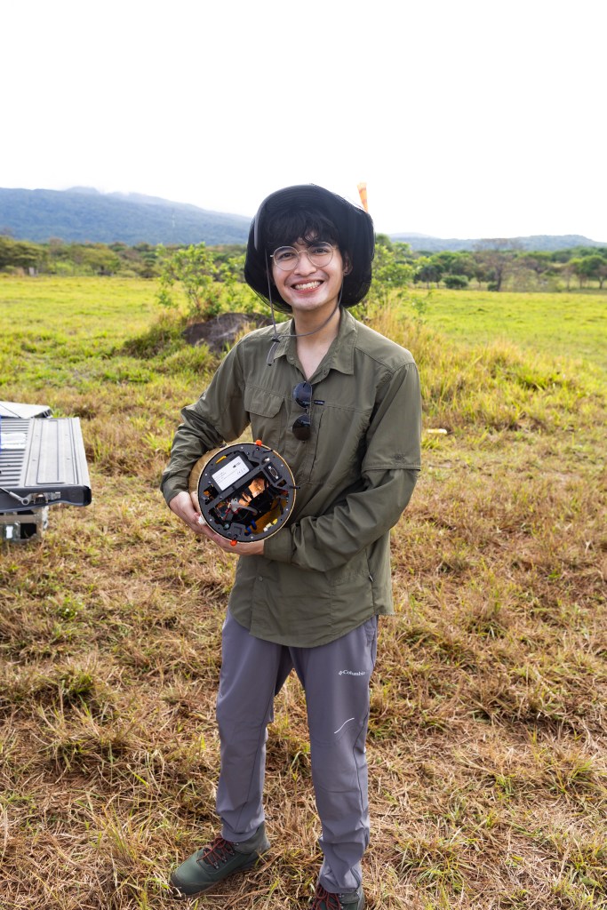 A man in an olive green longsleeve and black bucket hat smiles at the camera while holding a cylindrical device. He stands in a field of grass with a blue mountain visible in the background.