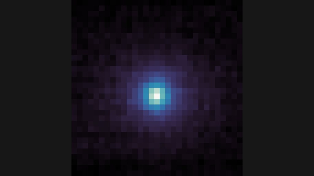 A square, pixelated image with a dark background. At the center is a roughly circular cluster of bright pixels, transitioning from white at the very center to light blue, then darker blue and purple as it spreads outward. The edges and corners of the image are mostly dark purple and black pixels.