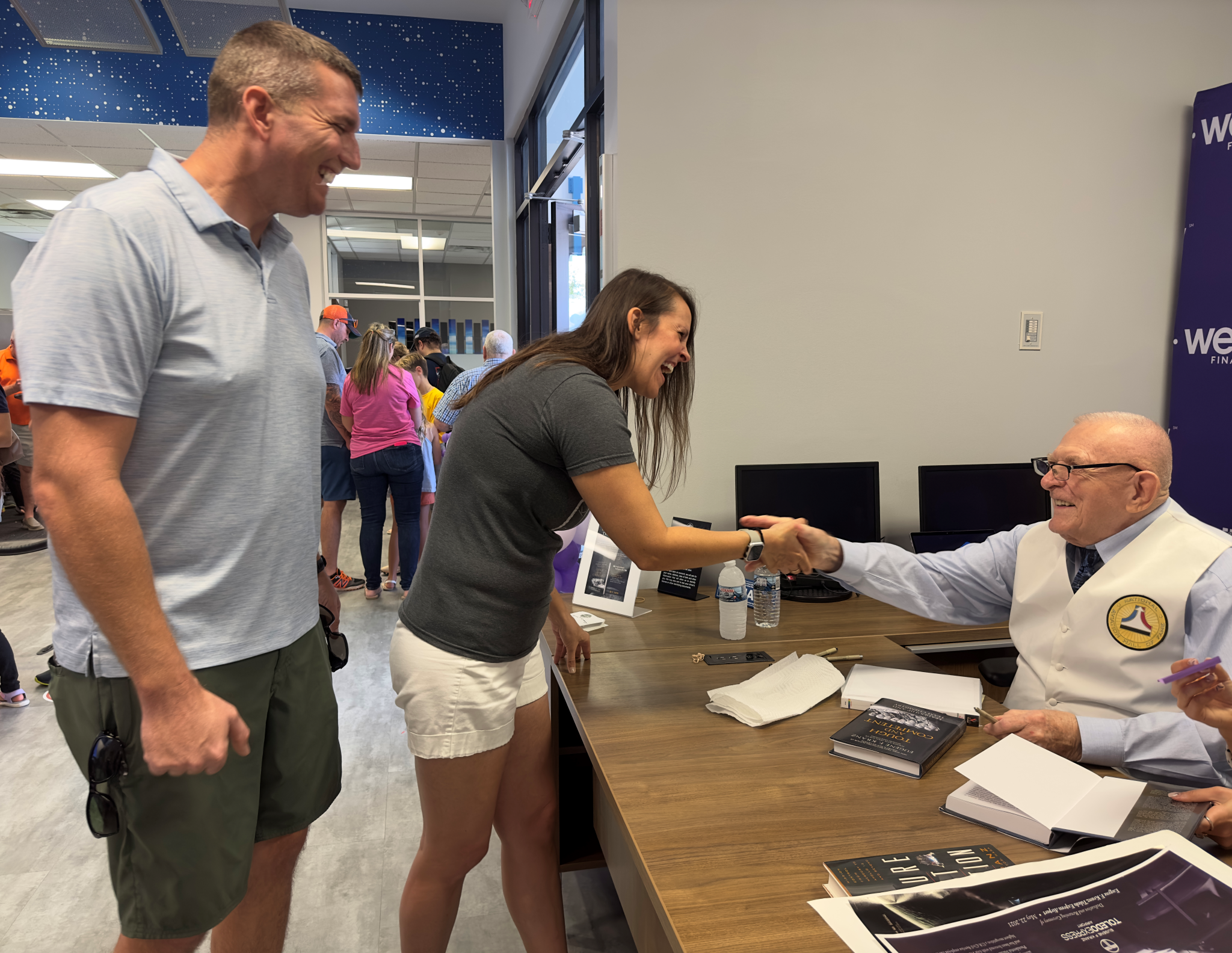 A man at a desk signs autographs. Two people stand in front of him as he shaked