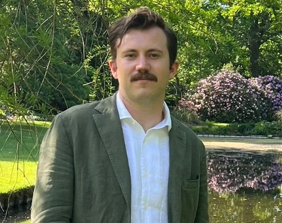 Portrait photo of NASA Ames scientist Aidan Ramey. He is standing in a green field wearing a white collared shirt.